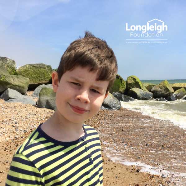 A depiction of Sarah's son,  young boy standing on top of a sandy beach with longleigh foundation's logo on the top right of the digital promotional asset.