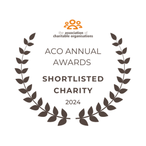 A circular emblem with a dark brown laurel wreath border. Inside the circle, the text reads: "ACO Annual Awards" in uppercase, followed by "Shortlisted Charity" in bold, larger font, and "2024" below. At the top, the logo of the Association of Charitable Organisations is displayed, featuring an abstract orange figure consisting of three interconnected circles with the organization's name below it in small text. The image has a transparent background.