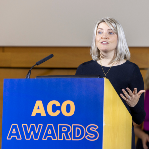Aileen Edmunds is a woman with blonde hair, dressed in a black dress, speaking at a podium during the ACO Awards. The podium is blue and yellow, displaying the words "ACO Awards" in bold orange lettering. She gestures with one hand while addressing the audience following Longleigh's win for the Collaboration of the Year Award, with a microphone positioned on the podium. Aileen is the CEO of Longleigh Foundation.