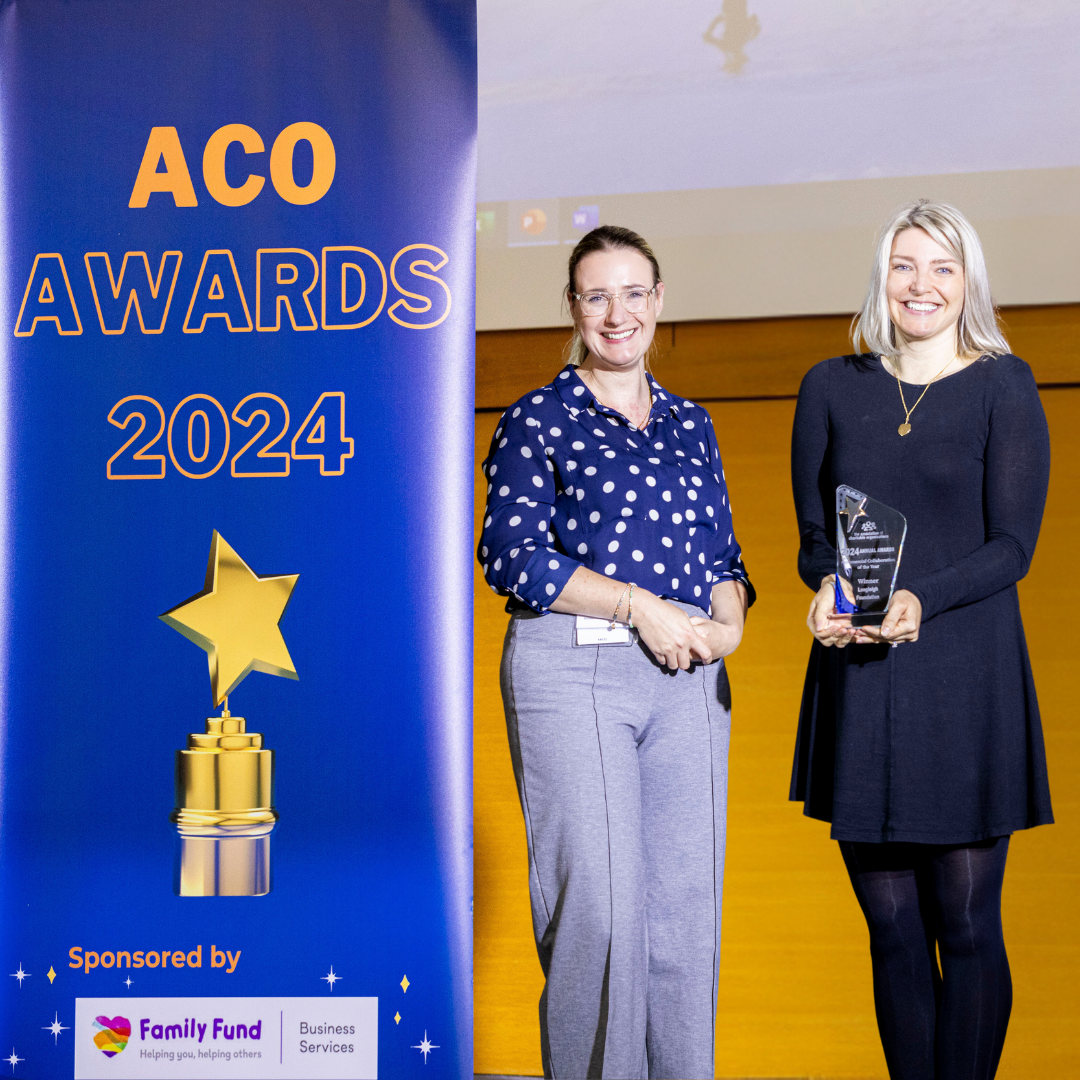 Two women stand on a stage, one holding a glass award trophy. They are beside a large blue banner that reads 