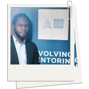 A photo of Dayne Donegan, who runs Evolving Mentoring. Dayne has short, curly hair and a beard, dressed in a light blue shirt and dark blazer, stands next to a blue backdrop featuring a white logo with a meditative figure and the letters "EM." The partial text "Evolving Mentoring" is visible behind him. The image is styled like a Polaroid photograph with a paper clip at the top right corner.