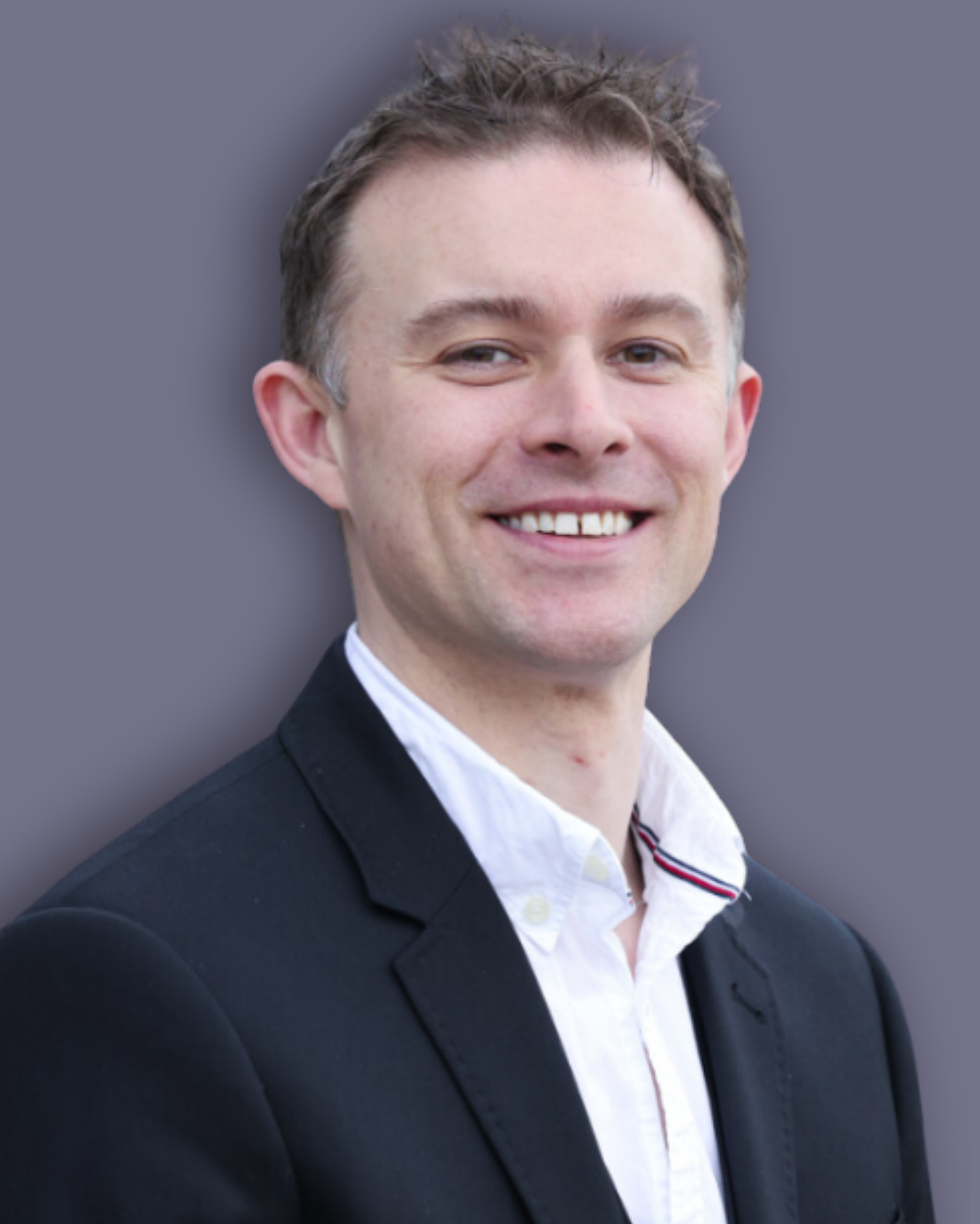 A headshot photo with a purple background of David Ripley who is wearing a black suit jacket over a white shirt. David has short blonde hair and has a bright smile.