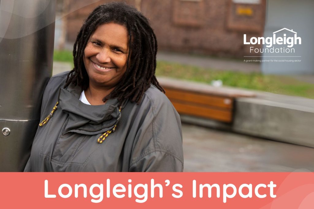 A woman depicting Jayne with black shoulder length dreadlocks and a warm smile leans against a metal post outdoors, wearing a dark grey jacket.  The Longleigh Foundation logo is displayed in the top right corner. The background is slightly blurred, showing a brick building and sidewalk. The lower part of the image shows the words 'Longleigh's Impact'